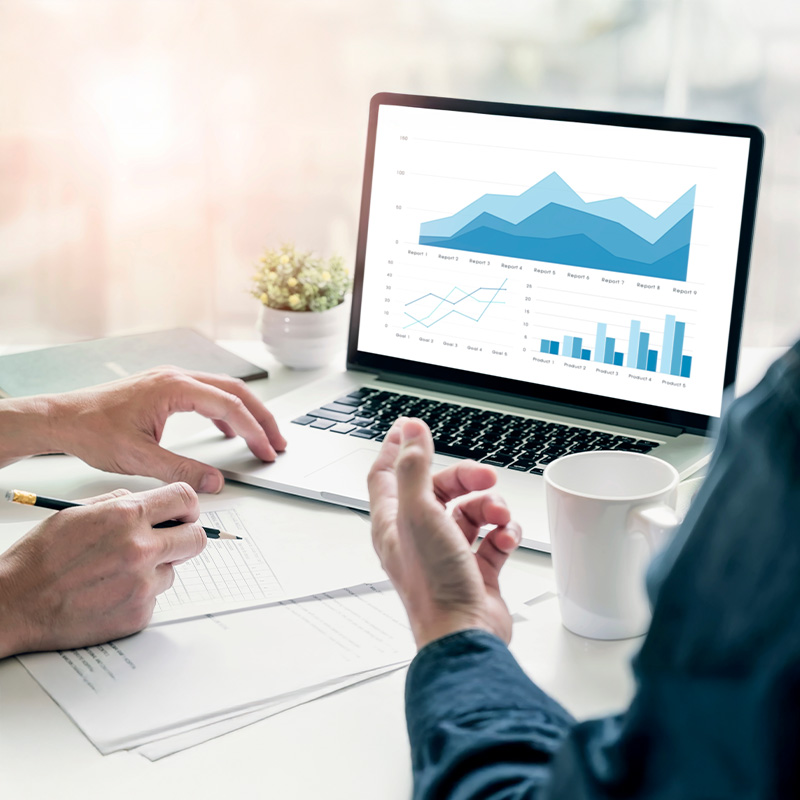 Dos personas revisan gráficos financieros y documentos sobre una mesa, mientras observan una pantalla de portátil con estadísticas y gráficos en tonos azules.