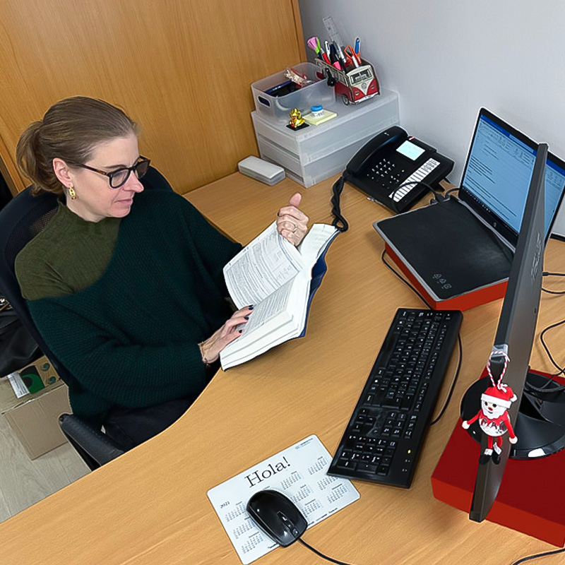 Una mujer sentada en un escritorio de oficina, leyendo un libro abierto. Frente a ella hay un ordenador con doble pantalla, teléfono, teclado y material de oficina.